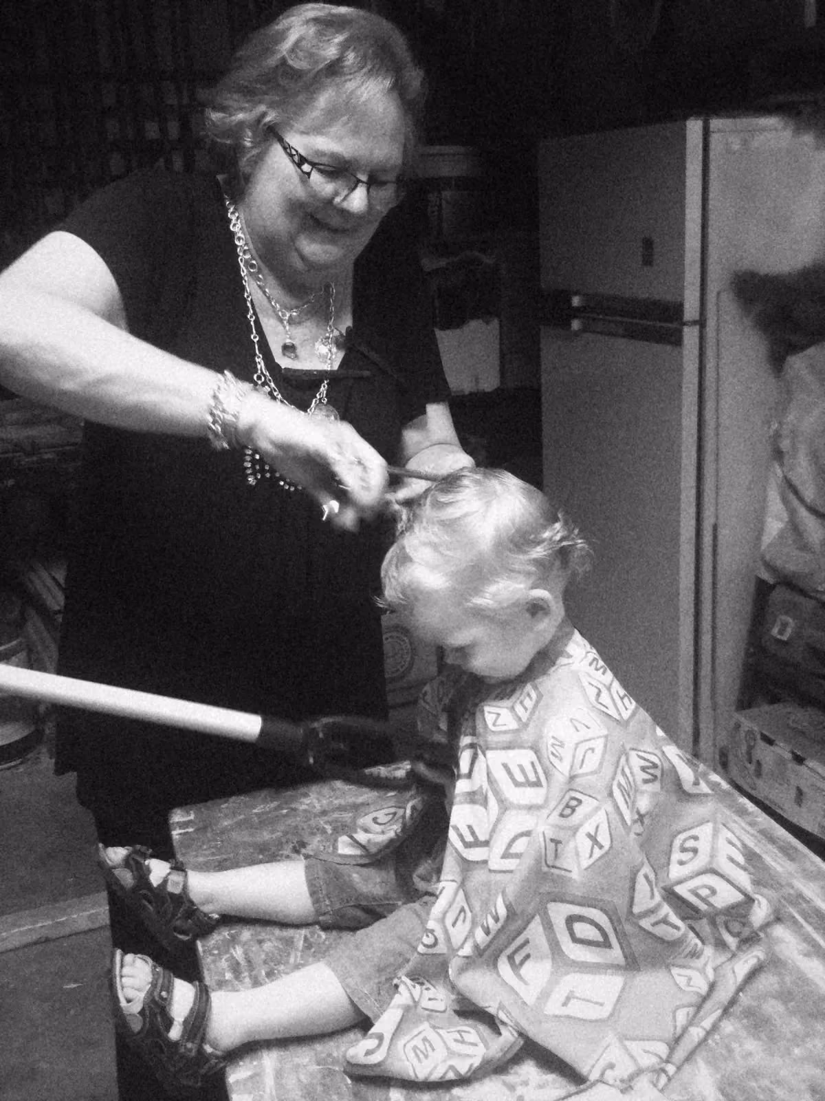 A toddler in an alphabet cape getting a haircut on a workbench out the back